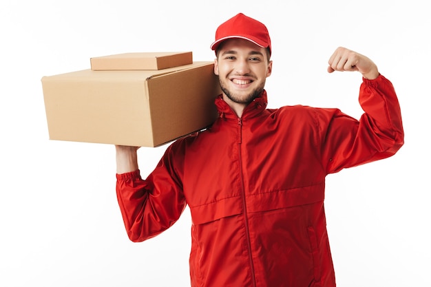 Repartidor sonriente joven en gorra roja y chaqueta sosteniendo el paquete en la mano mostrando felizmente la fuerza mientras