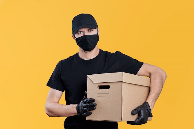 Repartidor en uniforme negro mantenga caja de cartón sobre fondo amarillo. Concepto de servicio de entrega sin contacto