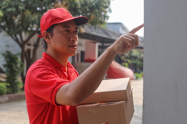 Repartidor con uniforme rojo y gorra sosteniendo una pila de cartón mientras toca el timbre de la casa