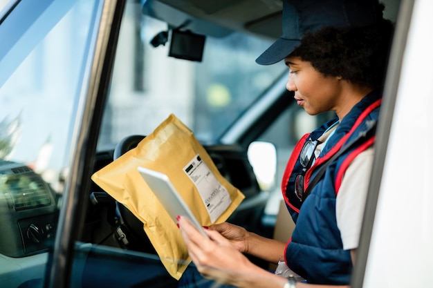 Repartidora afroamericana usando tableta digital mientras está sentada en una camioneta y revisando el paquete para la entrega