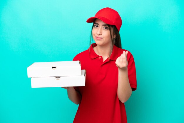 Repartidora de pizza con uniforme de trabajo recogiendo cajas de pizza aisladas de fondo azul haciendo gesto de dinero