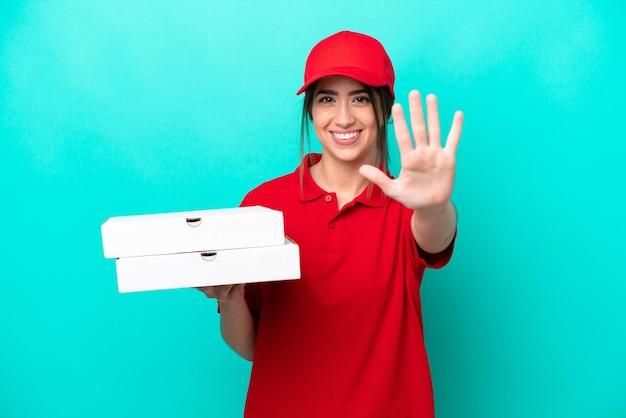 Foto repartidora de pizzas con uniforme de trabajo recogiendo cajas de pizza aisladas de fondo azul contando cinco con los dedos