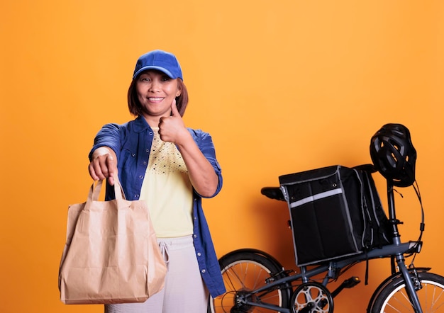 Una repartidora sonriente haciendo un gesto de aprobación mientras entrega comida para llevar a los clientes, usando bolsas de papel. trabajador de restaurante trayendo pedidos con bicicleta. servicio y concepto de comida para llevar