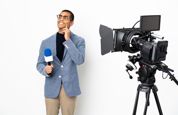 Reporter Afroamerikaner Mann, der ein Mikrofon hält und Nachrichten über isolierte weiße Wand berichtet, die eine Idee beim Aufschauen denken