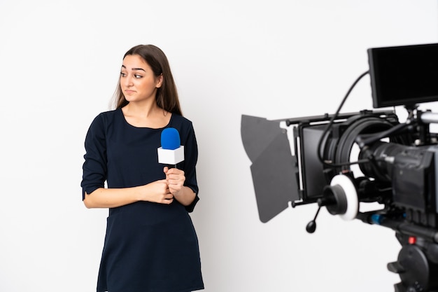 Repórter mulher segurando um microfone e reportando notícias isoladas na parede branca, fazendo dúvidas gesto olhando de lado