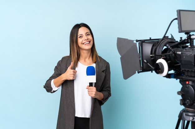 Foto repórter mulher segurando um microfone e reportar notícias sobre parede azul isolada, dando um polegar para cima gesto