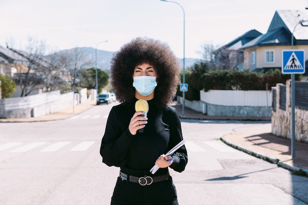 Reportero de televisión periodista con cabello afro, con una mascarilla quirúrgica para protegerse de un virus, informando sobre una historia en la calle