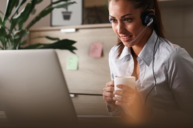 Foto representante de atendimento ao cliente sorridente com fone de ouvido usando um computador enquanto conversa com um cliente e bebe café no call center