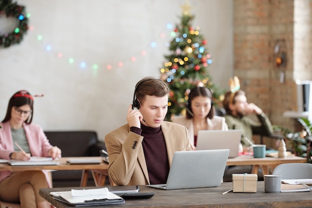 Representante de soporte al cliente masculino serio joven con auriculares mirando la pantalla de la computadora portátil mientras está sentado junto al escritorio y consultando a los clientes