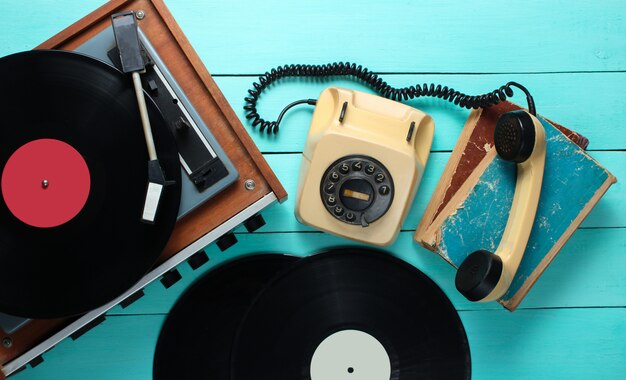 Reproductor de vinilo, teléfono rotativo, placas de vinilo, libros antiguos. Objetos anticuados en un fondo de madera azul. Estilo retro, años 70. Vista superior.