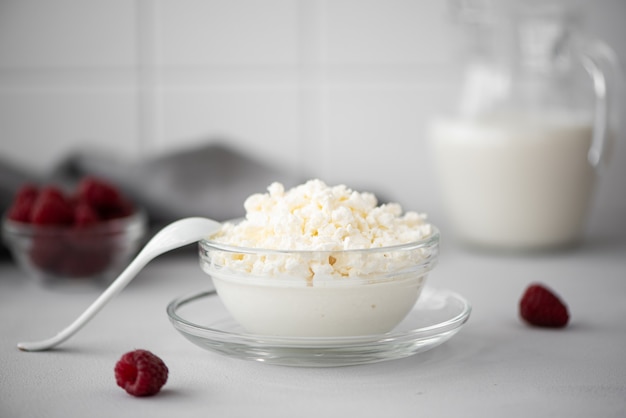 Requesón casero con leche y frambuesas en un recipiente de vidrio sobre una mesa blanca