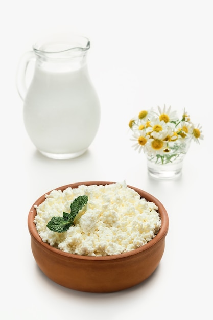 Requesón granulado en loza junto a una jarra de leche. Primer plano, enfoque selectivo, fondo blanco brillante. Requesón blando, comida sana natural, comida de dieta completa