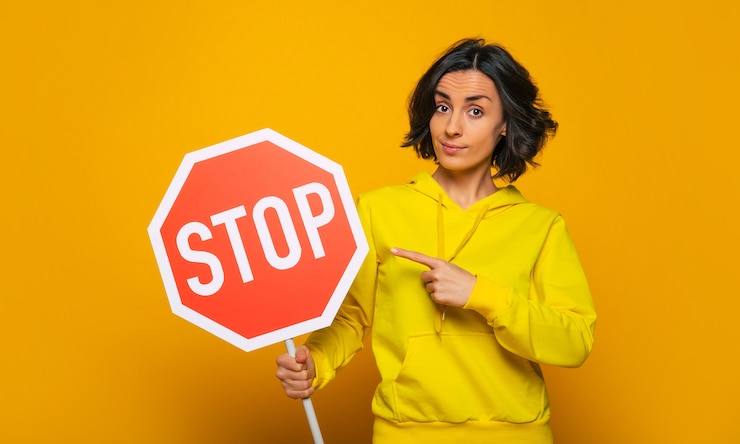 Foto requisito persistente. chica joven segura y persistente, vestida con una sudadera con capucha amarilla, señalando seriamente la señal de "pare" con el dedo.