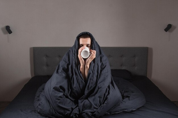 Resfriado y gripe Retrato de un joven enfermo bebiendo té en la cama caucásico enfermo sentado con una taza de bebida caliente en las manos con fiebre que no se siente bien en casa primer plano