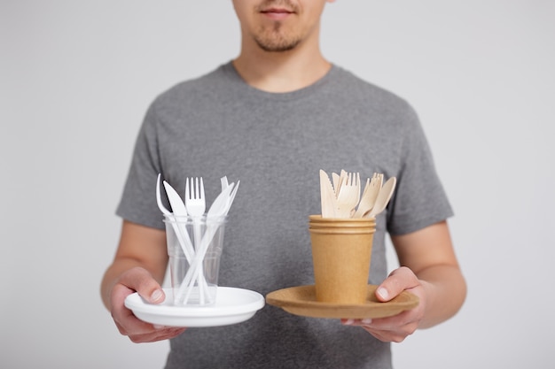 Resíduos zero e conceito ecológico - jovem comparando plástico com utensílios de mesa de madeira e papel sobre um fundo cinza