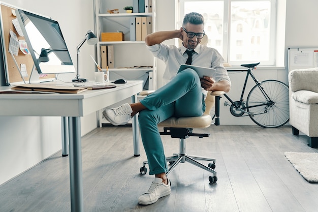 Resolviendo las cosas. Apuesto joven en camisa y corbata con tableta digital y sonriendo mientras está sentado en la oficina