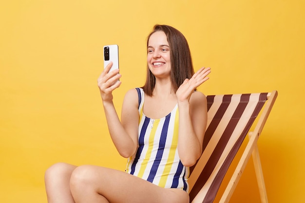 Resort-Blog Lächelnde Bloggerin in gestreiftem Badeanzug sitzt auf einem Liegestuhl isoliert vor gelbem Hintergrund und sendet einen Livestream aus dem Urlaub mit einem Videoanruf, der Hallo sagt