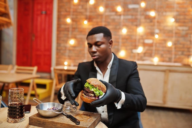 Respetable joven afroamericano con traje negro y guantes para comer sentado en un restaurante con sabrosa hamburguesa doble y bebida gaseosa