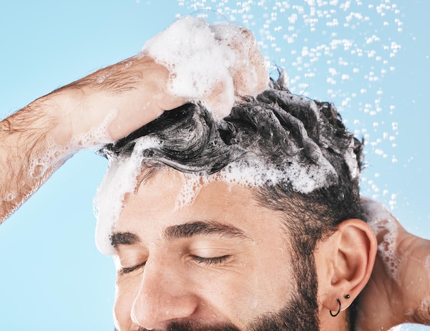 Foto respingos de água no rosto e homem no chuveiro com xampu no estúdio em um fundo azul gotas de água de dermatologia para cuidados com a pele e modelo masculino, banho ou lavagem para cuidados com os cabelos, higiene e bem-estar