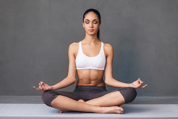 Respire fundo. Bela jovem africana em roupas esportivas, praticando ioga enquanto está sentado em posição de lótus contra um fundo cinza