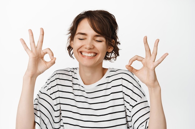 Respuesta positiva, excelente. Sonriente mujer feliz cierra los ojos, muestra signos de aprobación, me gusta y está de acuerdo, de pie en camiseta contra fondo blanco