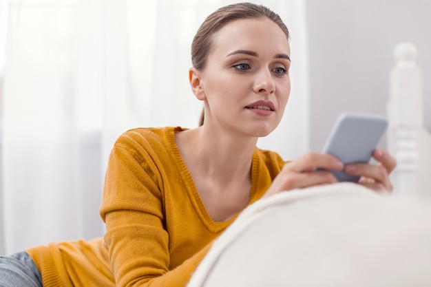 Respuesta urgente. Agradable mujer atractiva pensativa que usa el teléfono mientras mira la pantalla y posa en la luz