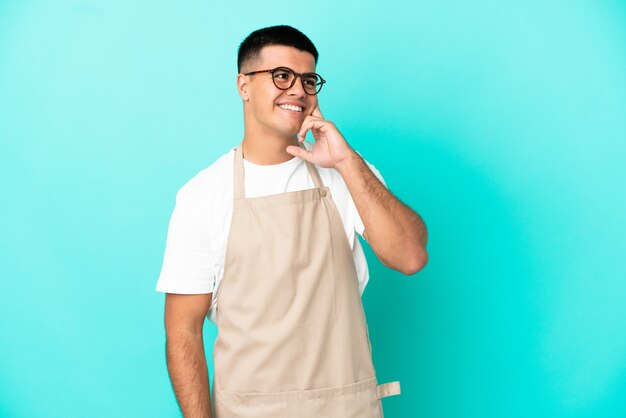 Restaurant-Kellner-Mann über isoliertem blauem Hintergrund, der eine Idee denkt, während er nach oben schaut