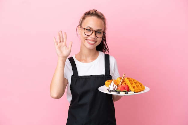 Restaurant Kellner russisches Mädchen mit Waffeln isoliert auf rosa Hintergrund grüßend mit der Hand mit glücklichem Ausdruck