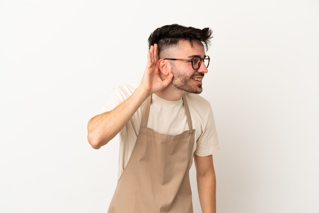 Restaurante camarero hombre caucásico aislado sobre fondo blanco escuchando algo poniendo la mano en la oreja