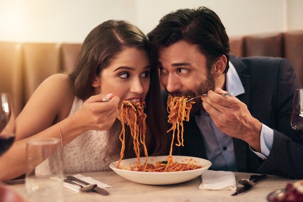 Restaurante faminto e casal comendo espaguete por amor, diversão louca e compartilhamento de prato na celebração do dia dos namorados Pessoas felizes com comida de macarrão para jantar de aniversário ou experiência gastronômica requintada