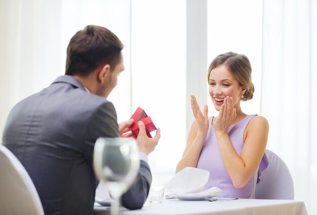 restaurante, pareja y concepto de vacaciones - joven emocionada mirando a su novio con una caja de regalo en el restaurante