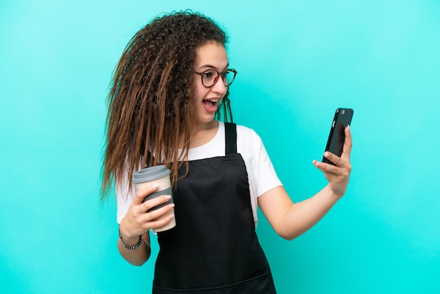 Restaurantkellner Arabische Frau isoliert auf blauem Hintergrund mit Kaffee zum Mitnehmen und einem Handy