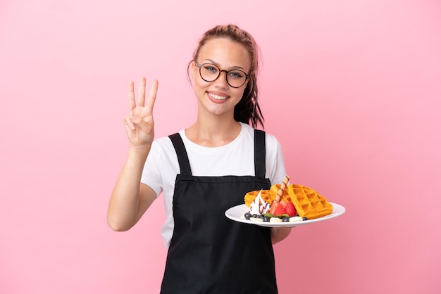 Restaurantkellner Russisches Mädchen hält Waffeln isoliert auf rosa Hintergrund glücklich und zählt drei mit den Fingern