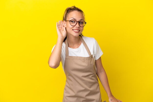 Restaurantkellner Russisches Mädchen isoliert auf gelbem Hintergrund, das etwas hört, indem es die Hand auf das Ohr legt