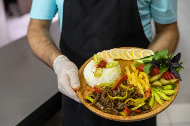 Restaurantkoch hält leckeres Fleischgericht mit Salat, Gemüse auf dem Holzteller