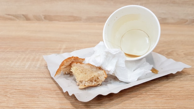 Restos de comida en un plato de papel y taza de café de papel sucio vacío en una mesa de madera en un café de comida rápida.