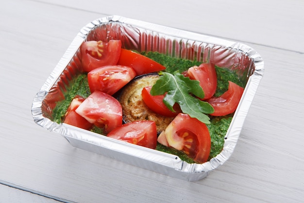 Retirar la comida en cajas de aluminio. Berenjena asada con guacamole y tomates frescos en madera blanca