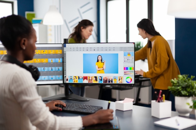 Retocador de mujer africana sentada en un estudio fotográfico creativo moderno trabajando en la computadora después de la sesión de fotos, retocando fotografías del modelo en el software de edición de imágenes con lápiz óptico, aplicación de degradado de color