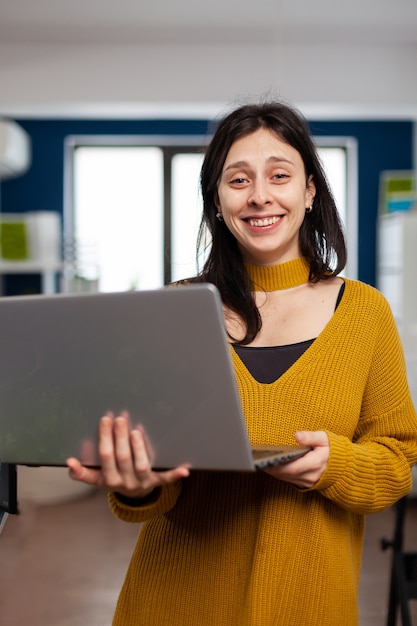 Retocador de mujer mirando a la cámara sonriendo trabajando en la agencia de medios creativos de pie en la empresa multimedia sosteniendo el portátil