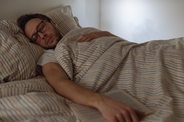 Foto retrato à noite de um homem adulto vestido com barba adormeceu com um livro na mão na cama antes de dormir