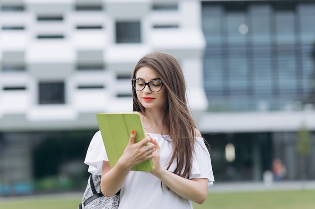 Retrato desde abajo de la joven y bella empresaria usando un dispositivo de tableta mientras está de pie al aire libre en un gran edificio urbano