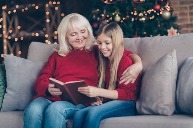 Retrato de abuela nieto sentarse en el diván disfrutar de leer el libro de cuento de hadas en la habitación decorada de la casa loft