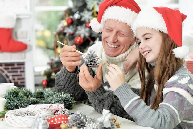 Retrato de abuelo con nieta en gorros de Papá Noel preparándose para Navidad en casa