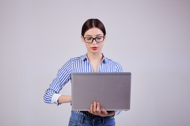 Retrato de una administradora en una camisa blanca y azul a rayas con gafas y una computadora portátil en gris. Empleada del año, mujer de negocios. Ocupado.