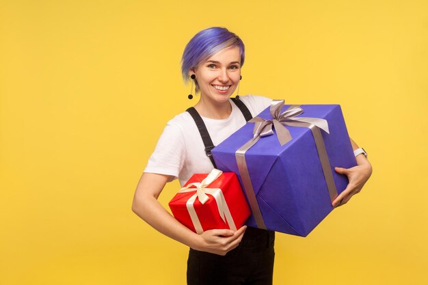 Retrato de una admirada y alegre hipster con el pelo corto violeta en overoles sosteniendo dos regalos, grandes y pequeños, cajas de regalo de vacaciones mirando a la cámara con una sonrisa dentuda, una foto de estudio de fondo amarillo