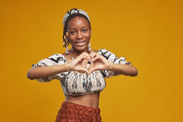 Retrato de una adolescente africana haciendo forma de corazón con las manos y sonriendo a la cámara de pie