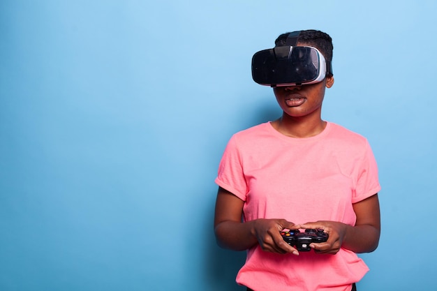 Retrato de adolescente afroamericano con casco de realidad virtual con controlador de juegos jugando videojuegos de shoother de espacio en línea en estudio con fondo azul. Concepto de videojuegos