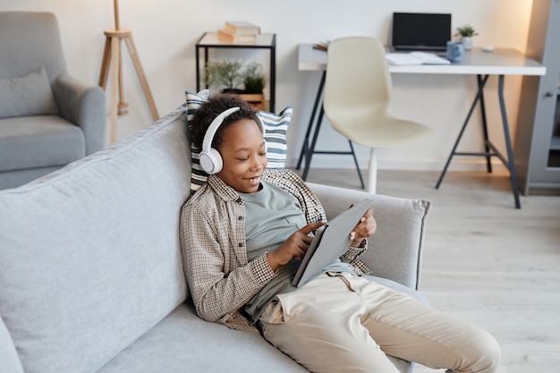 Retrato de adolescente afroamericano con tableta digital con auriculares mientras se relaja en el sofá en casa, espacio de copia