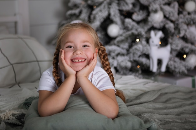 Retrato de una adolescente con coletas acostada en una cama con almohadas en el fondo de un árbol de Navidad. Enfoque selectivo, fondo borroso.