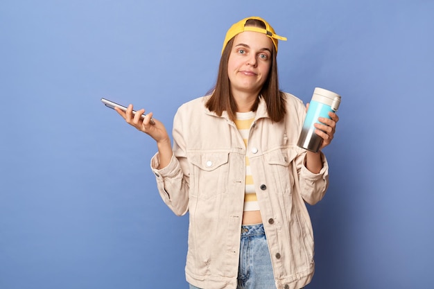 Retrato de una adolescente confundida e incierta con gorra de béisbol y chaqueta posando aislada sobre fondo azul sosteniendo un teléfono móvil que no sabe qué responder al mensaje de lectura